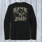 A black long-sleeve t-shirt with a large back graphic featuring a hawk and the words 'HAWK AND DAD' in distressed beige and green lettering, displayed against a light gray wood plank background.