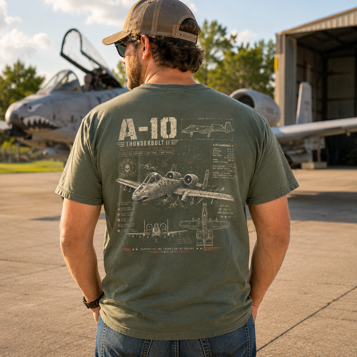A man viewed from behind wearing an olive green t-shirt with a detailed A-10 Thunderbolt II aircraft graphic on the back, standing on an airfield with military aircraft and a hangar in the background.