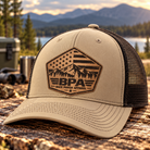 Richardson 112FP khaki and black mesh trucker hat with light tan BPA American flag mountain leather patch, featuring pine trees and Back Porch Apparel logo.