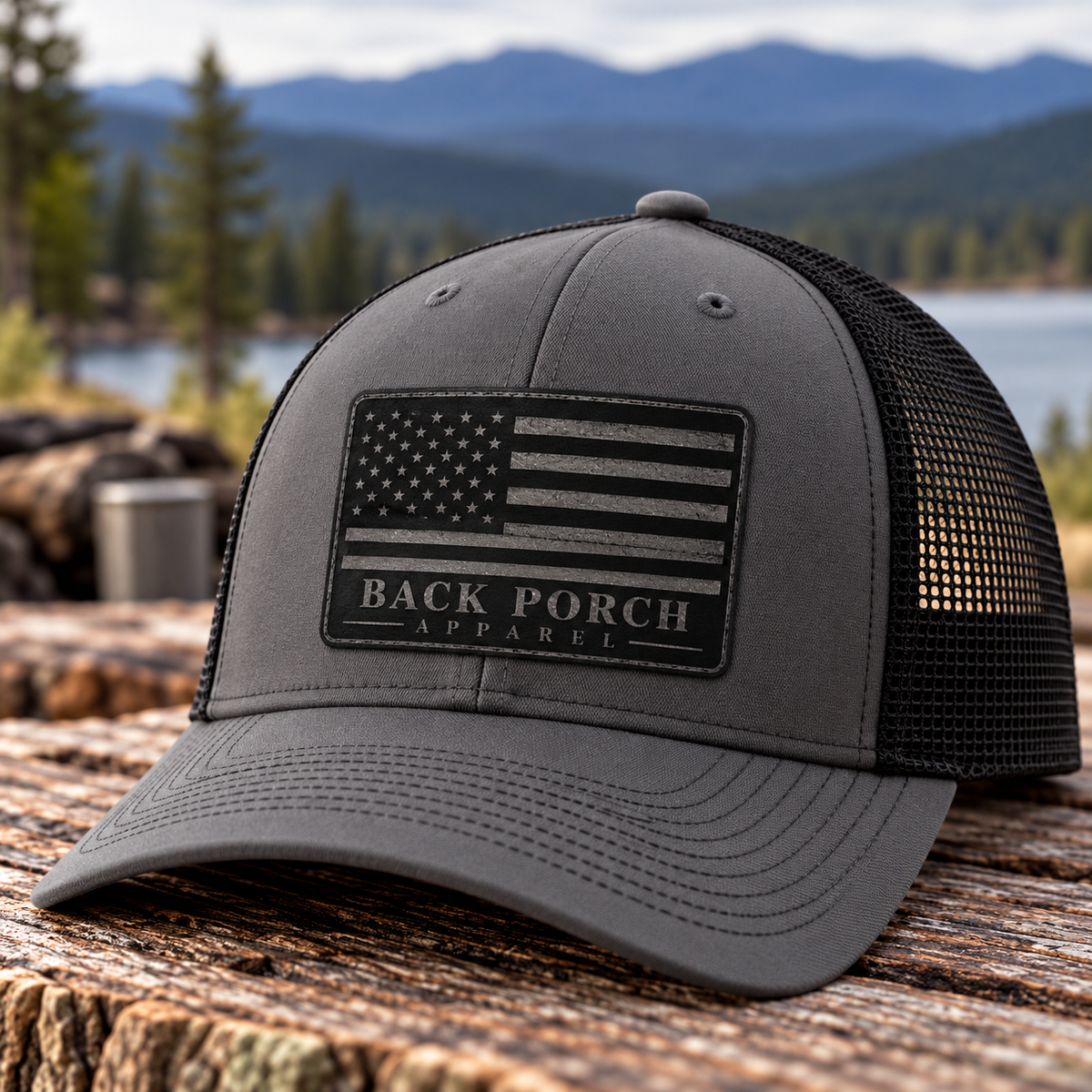 Gray and black Richardson five-panel trucker hat with black mesh back and charcoal leather patch featuring an American flag and Back Porch Apparel logo, displayed outdoors on rustic wood with mountains in the background.