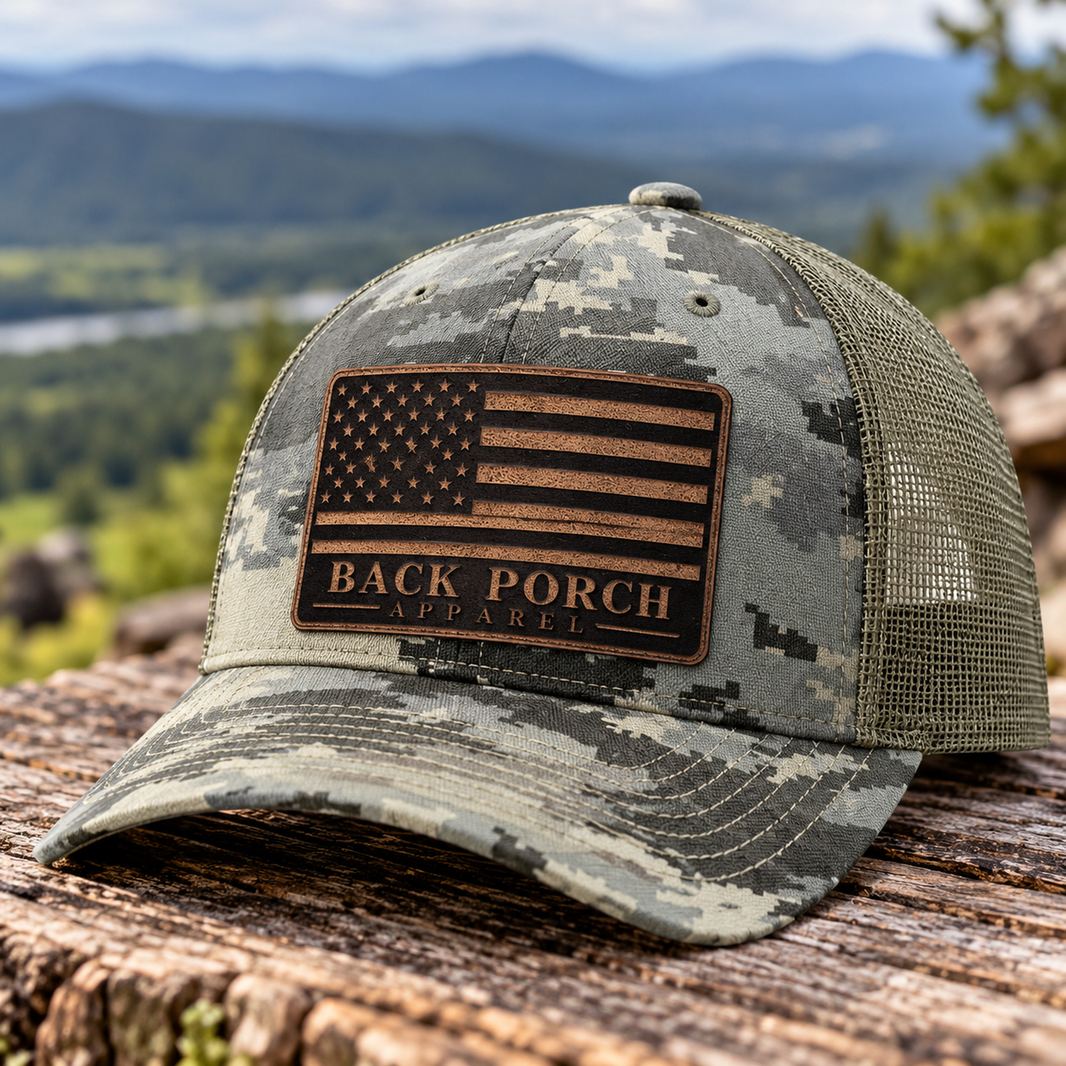 Richardson 112PFP digital camouflage trucker hat with light green mesh back and tan leather patch featuring an American flag and Back Porch Apparel logo, displayed on rustic wood with a scenic mountain backdrop.