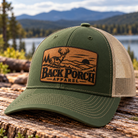 Richardson 112FP Loden green trucker hat with tan mesh back and light tan leather patch featuring a whitetail buck, mountain landscape, pine trees, and Back Porch Apparel logo, displayed on rustic wood with a scenic mountain lake background.