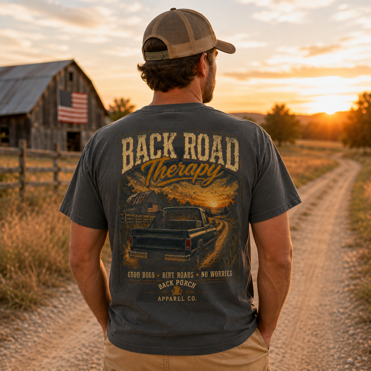 A man stands on a dirt road at sunset wearing a dark charcoal t-shirt with a large vintage-style back graphic that reads 'BACK ROAD Therapy' and shows a pickup truck on a country road. The shirt also includes small text reading 'GOOD DOGS - BENT BEARS - NO WORRIES' and 'SACI POPSY APPAREL CO.
