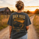 A woman stands on a dirt road at sunset wearing a dark charcoal graphic T-shirt with a large back print reading “BACK ROAD Therapy” and showing a vintage pickup truck scene. A barn and open field are visible in the background.