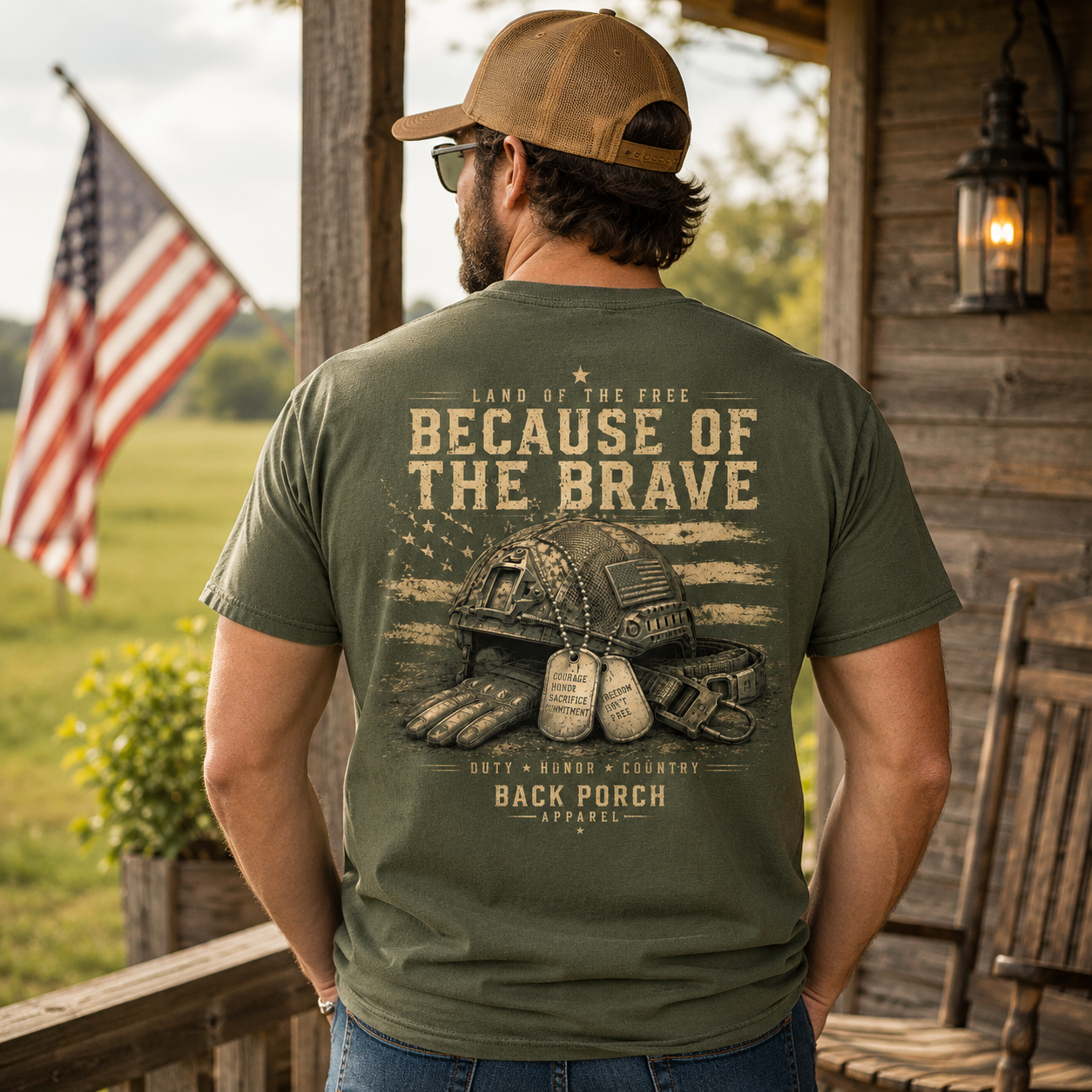 A man stands on a wooden porch wearing an olive green t-shirt with a large military-themed graphic on the back. The graphic features a military helmet, dog tags, and the text 'LAND OF THE FREE BECAUSE OF THE BRAVE' and 'DUTY HONOR COUNTRY BACK PORCH APPAREL'.