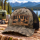 Bigfoot Believe leather patch trucker hat by Back Porch Apparel, featuring a light tan and black Sasquatch mountain design on a woodland camouflage Richardson trucker cap with black mesh back.