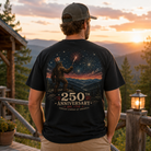 A man stands on a wooden porch at sunset wearing a black graphic T-shirt. The back of the shirt features a large printed design of a Bigfoot figure under fireworks with the text '250th Anniversary' and 'Est.