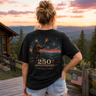 A woman stands outdoors at sunset wearing a black graphic T-shirt from the back, showing a large print of a Bigfoot figure under fireworks with the text '250th Anniversary' and 'United States of America.'.