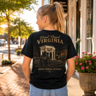 A woman viewed from behind wearing a black graphic t-shirt with a vintage-style illustration and text reading 'Front Royal, VIRGINIA' and 'BACK PORCH APPAREL', standing on a sunlit brick sidewalk in front of storefronts.