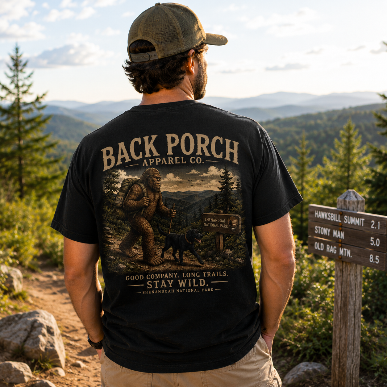 A man standing outdoors in a forest with mountains in the background, wearing a black graphic t-shirt from the brand Back Porch Apparel Co. The back of the shirt features a large vintage-style illustration of a Bigfoot scene with the text 'BACK PORCH APPAREL CO.