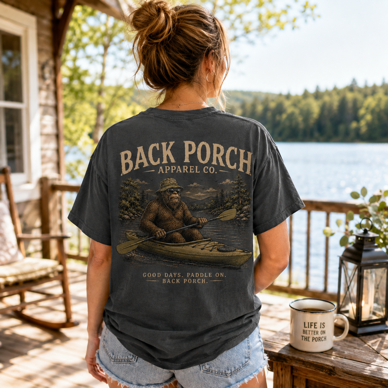 A woman stands on a porch by a lake, shown from the back, wearing a dark charcoal graphic T-shirt with a large vintage-style print of a bear paddling in a canoe and the text 'BACK PORCH APPAREL CO.' and 'GOOD DAYS, FADDLE ON, BACK PORCH.