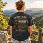 A woman stands outdoors on a mountain overlook wearing a dark charcoal graphic T-shirt with a large back print reading 'SKYLINE DRIVE VIRGINIA' and 'BACK PORCH APPAREL CO.' above and below an illustrated mountain scene.