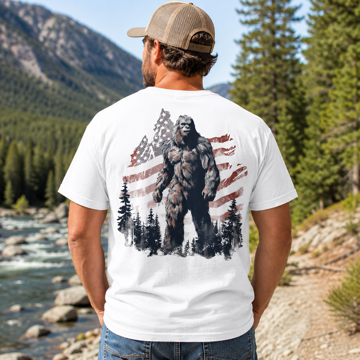 A man standing outdoors in a mountain forest setting, viewed from behind, wearing a white short-sleeve T-shirt with a large graphic print of Bigfoot in front of an American flag and pine trees.
