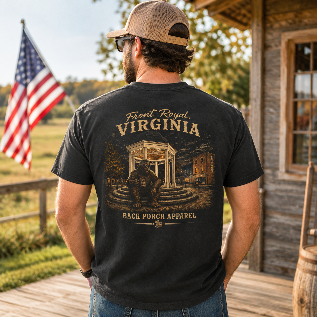 A man wearing a black graphic t-shirt with a vintage-style Virginia front porch design, shown from the back. He is standing on a wooden porch in front of a rustic cabin with an American flag in the background.