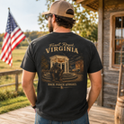 A man wearing a black graphic t-shirt with a vintage-style Virginia front porch design, shown from the back. He is standing on a wooden porch in front of a rustic cabin with an American flag in the background.