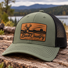 An olive green and black trucker hat with a brown leather patch on the front, set outdoors on a log with a lake and mountains in the background. The patch reads 'Buck Country - Back Porch Apparel'.