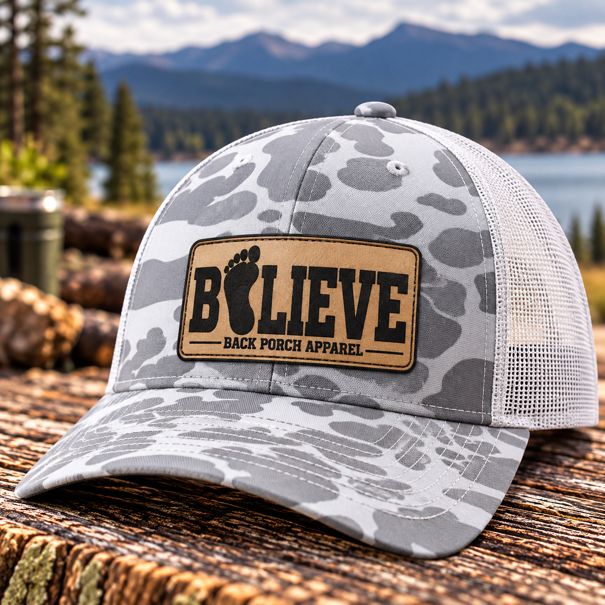 Richardson 112PFP Blizzard Duck Camo trucker hat with white mesh back and light tan BELIEVE Bigfoot footprint leather patch by Back Porch Apparel, photographed on rustic wood with a scenic mountain lake background.