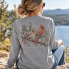 A woman sitting outdoors on a rock by a lake, seen from behind, wearing a heather gray long-sleeve shirt with a large back graphic of two cardinals on branches. The text 'SILK PITCH APPAREL' is printed below the graphic.
