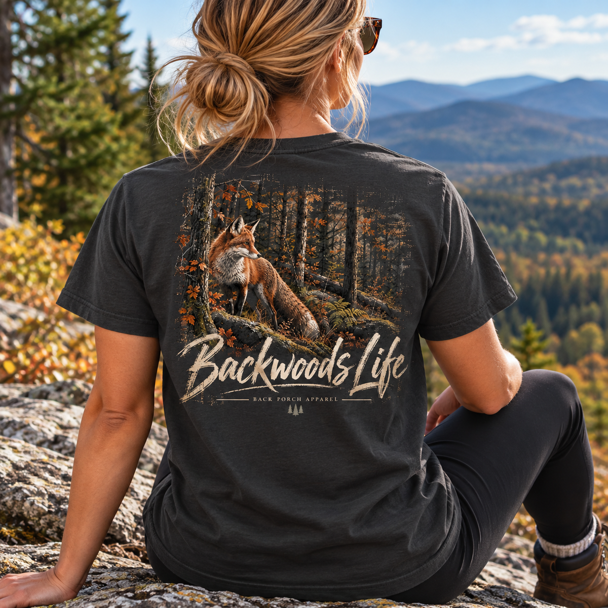 A woman sits outdoors on a rocky hillside with mountains and autumn trees in the background, wearing a dark charcoal T-shirt from the back. The shirt features a large graphic of a fox in a forest with the text 'Backwoods Life' and smaller text 'EACH TREE AFFAIR'.
