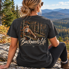 A woman sits outdoors on a rocky hillside with mountains and autumn trees in the background, wearing a dark charcoal T-shirt from the back. The shirt features a large graphic of a fox in a forest with the text 'Backwoods Life' and smaller text 'EACH TREE AFFAIR'.