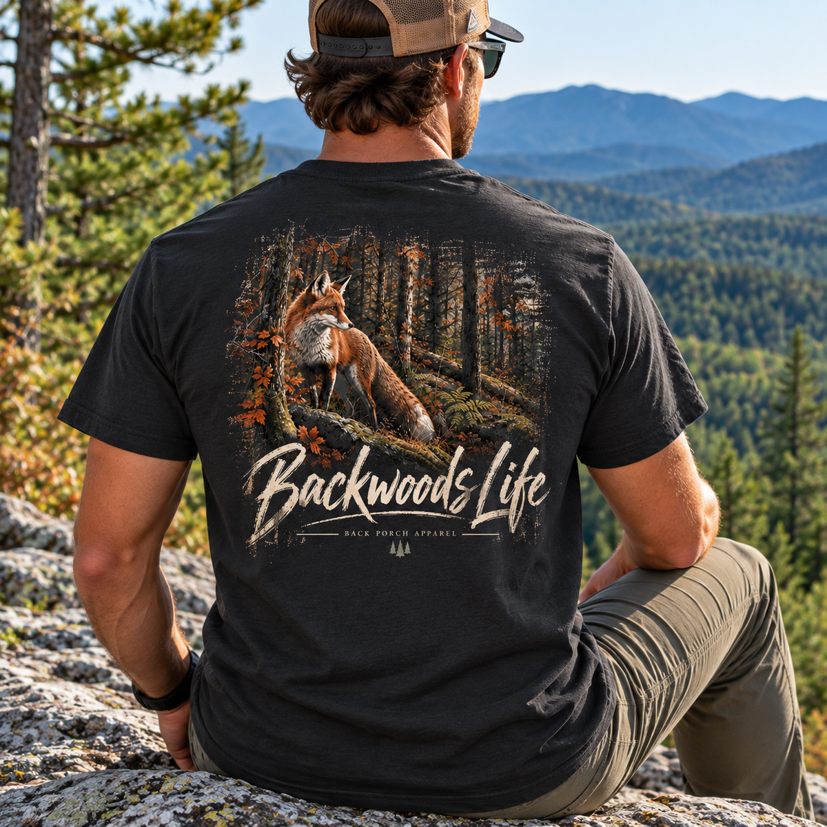 A man sitting outdoors on a rocky mountain overlook, viewed from behind, wearing a black graphic t-shirt with a large back print of a fox in a forest and the text 'Backwoods Life'.