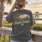 A woman viewed from behind wearing a dark charcoal gray graphic t-shirt with a vintage-style South Carolina landscape print on the back, standing outdoors near a wooden railing and water at sunset.