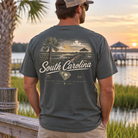A man viewed from behind wearing a dark gray t-shirt with a vintage-style South Carolina landscape graphic on the back, standing outdoors near a lake at sunset.