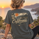 A woman sits outdoors by the ocean at sunset wearing a dark charcoal gray t-shirt with a large vintage-style California graphic on the back, featuring palm trees, mountains, an orange sun, and the words 'California' and 'The Golden State'.