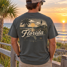 A man stands outdoors at sunset wearing a dark charcoal gray t-shirt with a large graphic print on the back featuring a Florida lighthouse scene and the word 'Florida' in cream lettering.