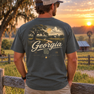 A man stands outdoors at sunset wearing a dark gray t-shirt with a large vintage-style Georgia landscape graphic on the back, paired with khaki pants and a baseball cap.