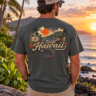 A man stands outdoors on a beach at sunset wearing a dark charcoal gray t-shirt with a large vintage-style Hawaii graphic on the back, paired with khaki pants and a baseball cap.
