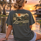 A woman sitting on a wooden dock at sunset wearing a dark gray t-shirt with a vintage-style Louisiana graphic on the back, with a lake and house in the background.