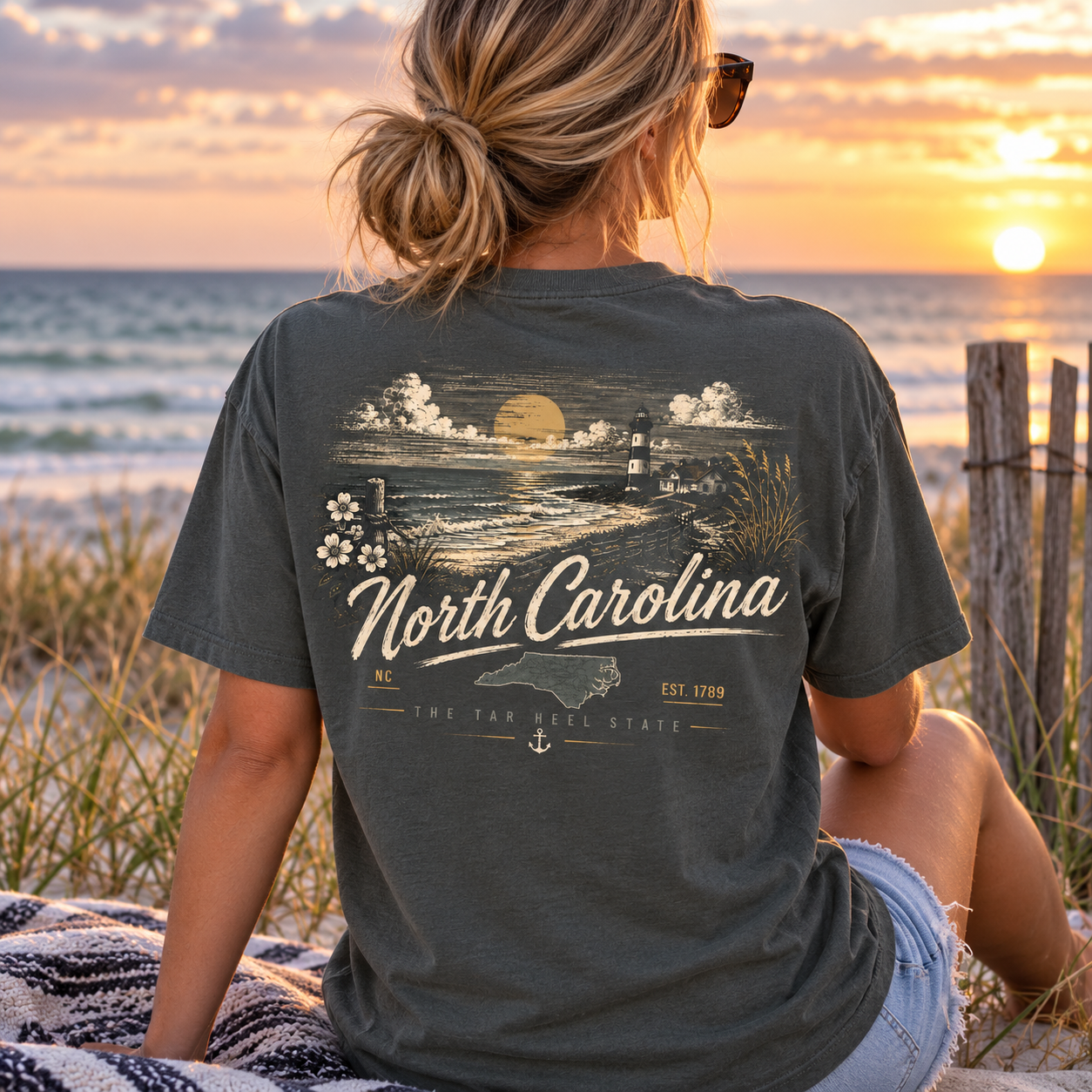 A woman sits on a beach at sunset wearing a dark charcoal gray t-shirt with a vintage-style 'North Carolina' graphic on the back, paired with denim shorts.