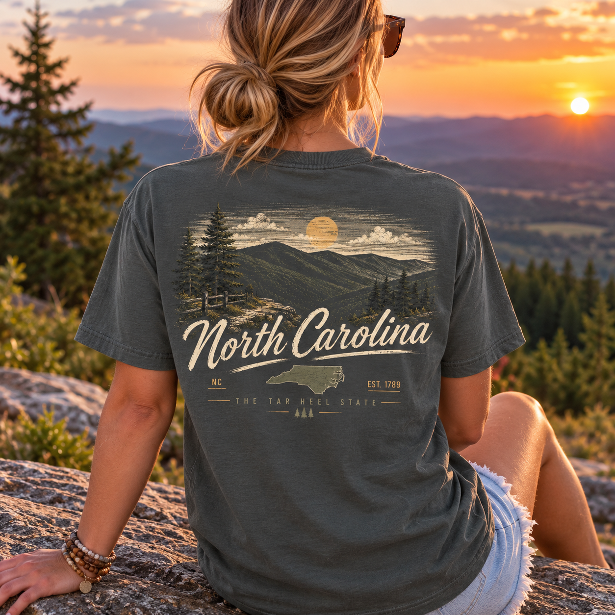 A woman sitting outdoors on a rock at sunset wearing a dark charcoal gray t-shirt with a vintage-style North Carolina mountain landscape graphic on the back.