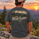A man viewed from behind wearing a dark gray t-shirt with a detailed North Carolina mountain landscape graphic on the back, set against a scenic sunset mountain background.