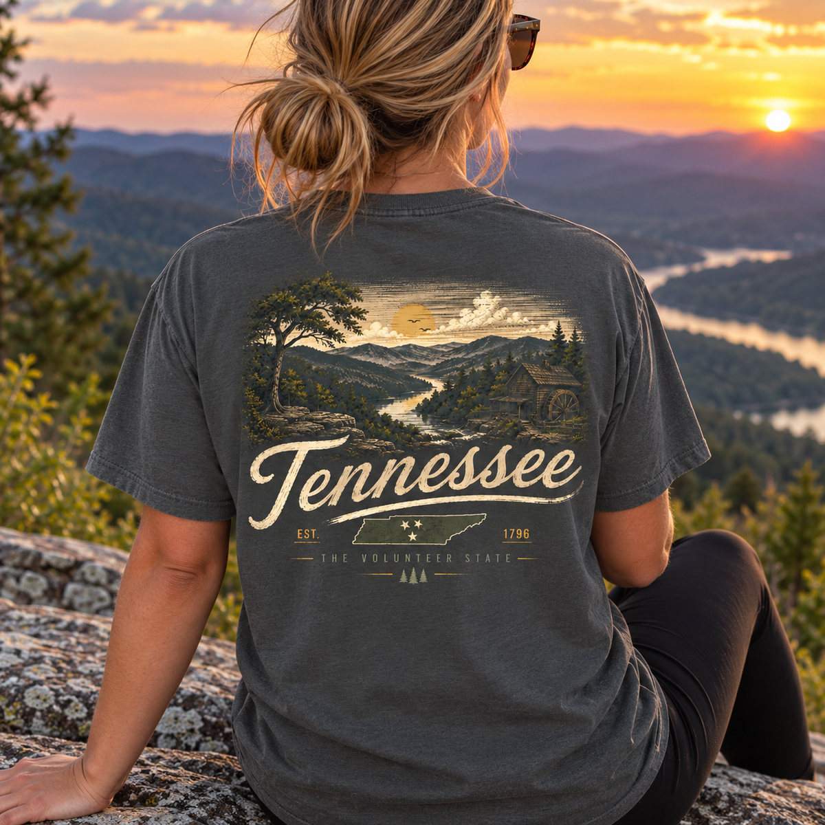 A woman sits on a rock overlooking a mountain landscape at sunset, wearing a dark charcoal gray t-shirt with a large vintage-style Tennessee graphic on the back.