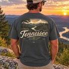 A man viewed from behind wearing a dark charcoal gray t-shirt with a large graphic print on the back showing a Tennessee landscape and the text 'Tennessee'. The background is a scenic mountain sunset with a river.