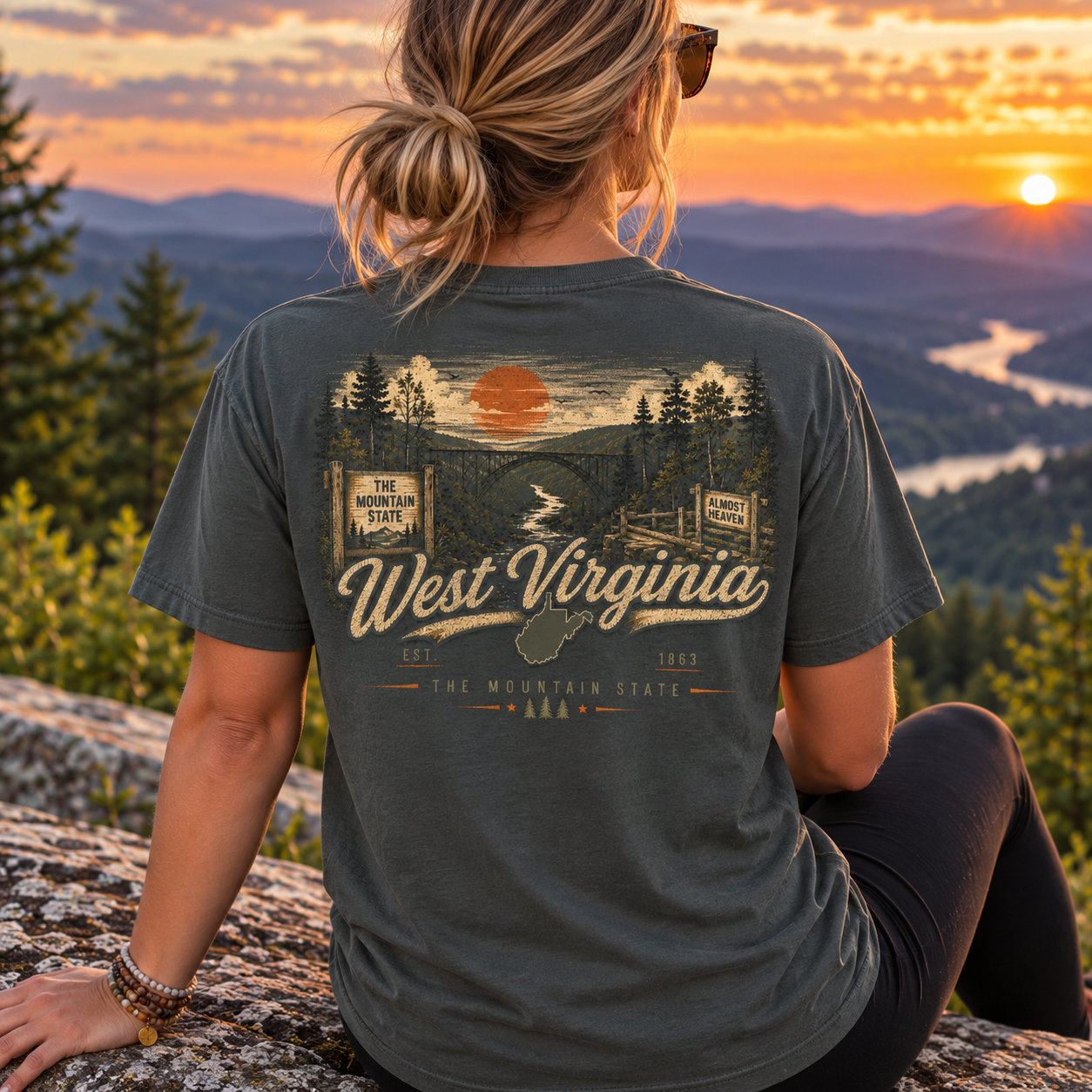 A woman sits on a rock outdoors at sunset wearing a dark charcoal gray graphic T-shirt. The back of the shirt features a scenic mountain landscape print with the words 'West Virginia' and 'The Mountain State.