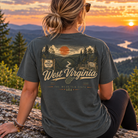A woman sits on a rock outdoors at sunset wearing a dark charcoal gray graphic T-shirt. The back of the shirt features a scenic mountain landscape print with the words 'West Virginia' and 'The Mountain State.
