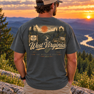 A man viewed from behind wearing a dark heather gray t-shirt with a large graphic print of 'West Virginia' and a mountain landscape at sunset. He is standing outdoors on a rocky hill overlooking a scenic river valley at sunset.