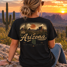 A woman sits outdoors at sunset wearing a black graphic T-shirt with a desert landscape print and the word Arizona across the back, paired with blue jeans.