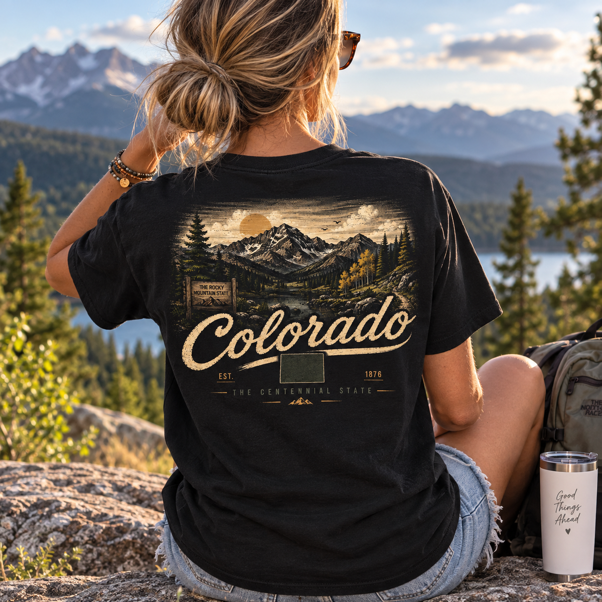 A woman sitting outdoors on a rock in front of a mountain and lake landscape, wearing a black graphic T-shirt with a large Colorado mountain scene and the word Colorado across the back.
