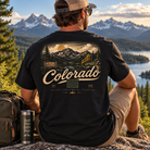 A man sits outdoors on a rock overlooking a mountain lake landscape, wearing a black graphic T-shirt with a large Colorado mountain scene and the word Colorado across the back. A backpack and insulated tumbler are beside him.