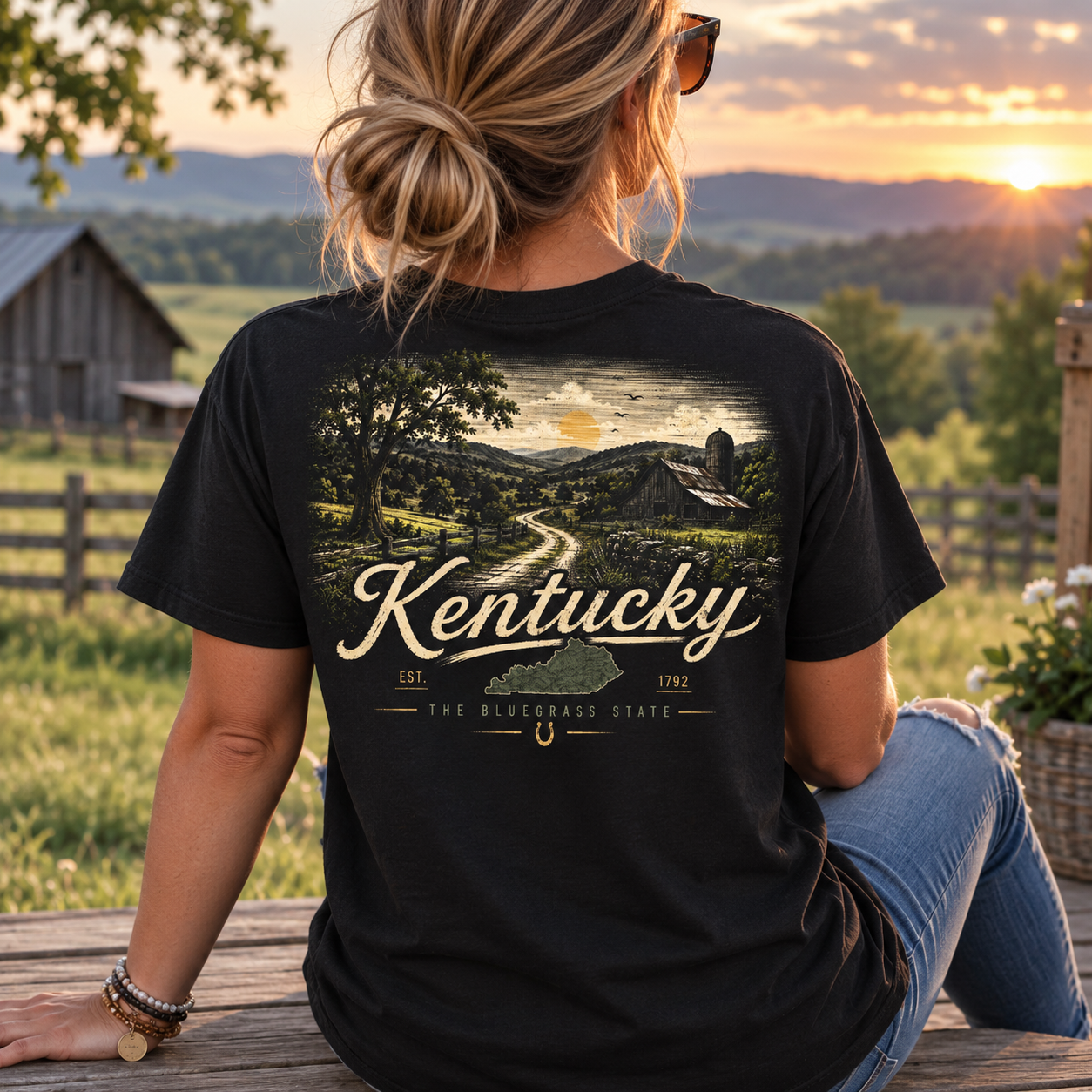 A woman sits on a wooden fence outdoors at sunset wearing a black graphic T-shirt. The back of the shirt features a scenic landscape print with the word 'Kentucky' in large cream lettering and smaller text reading 'The Bluegrass State' plus 'Est.
