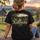A woman sits on a wooden fence outdoors at sunset wearing a black graphic T-shirt. The back of the shirt features a scenic landscape print with the word 'Kentucky' in large cream lettering and smaller text reading 'The Bluegrass State' plus 'Est.