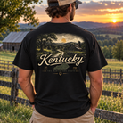 A man stands outdoors at sunset wearing a black graphic T-shirt. The back of the shirt features a large vintage-style Kentucky landscape print with a road, trees, hills, and the word 'Kentucky' in cream lettering.