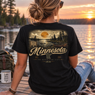 A woman sits on a wooden dock at sunset wearing a black graphic T-shirt with a large Minnesota-themed outdoor scene printed on the back. The design includes a lake, trees, a canoe, and the words 'Minnesota' and 'The North Star State.