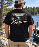 A man stands outdoors by a lake wearing a black graphic T-shirt from the back. The shirt features a scenic Minnesota lakeside print with a canoe, trees, and large white 'Minnesota' lettering.