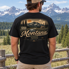 A man viewed from behind wearing a black graphic t-shirt with a vintage Montana landscape print on the back, set against a background of mountains and a wooden fence.