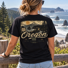 A woman viewed from behind wearing a black graphic t-shirt with a vintage-style Oregon landscape print, standing on a wooden railing with a scenic ocean coastline in the background.